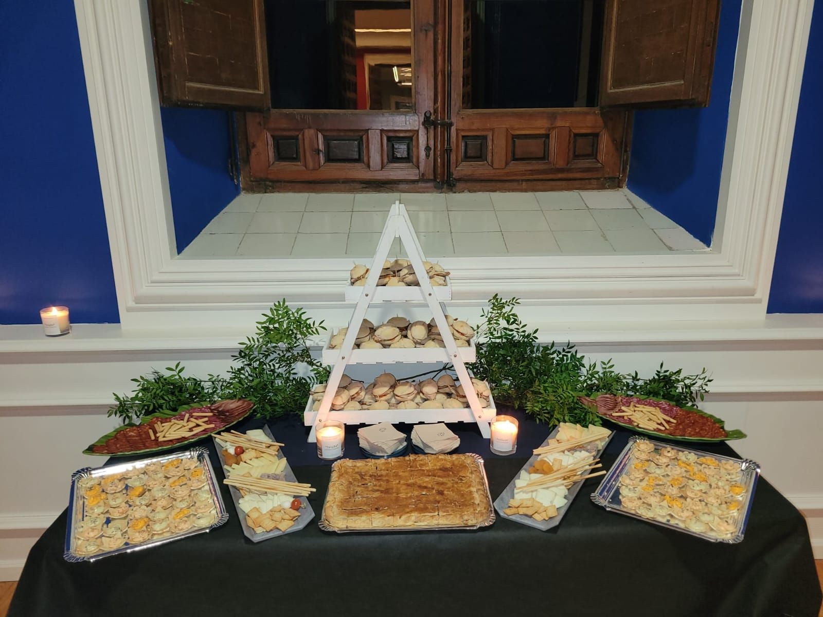 Exhibición de comida sobre un mantel negro, con pastel, sándwiches y pasteles. Estantes blancos muestran más comida. Pared azul con contraventanas de madera.