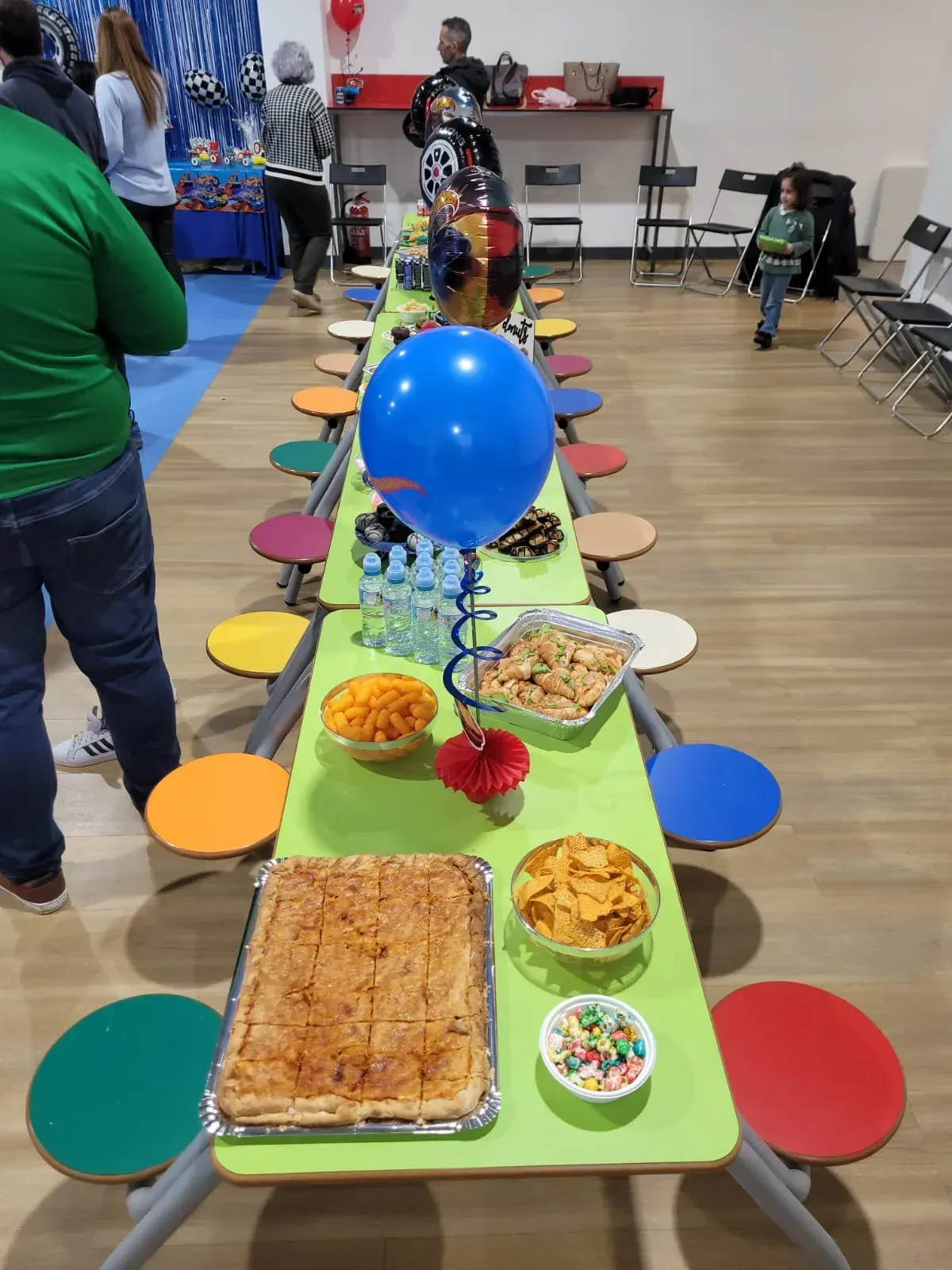 Mesa larga con comida y taburetes de colores, globo azul, gente de fondo.