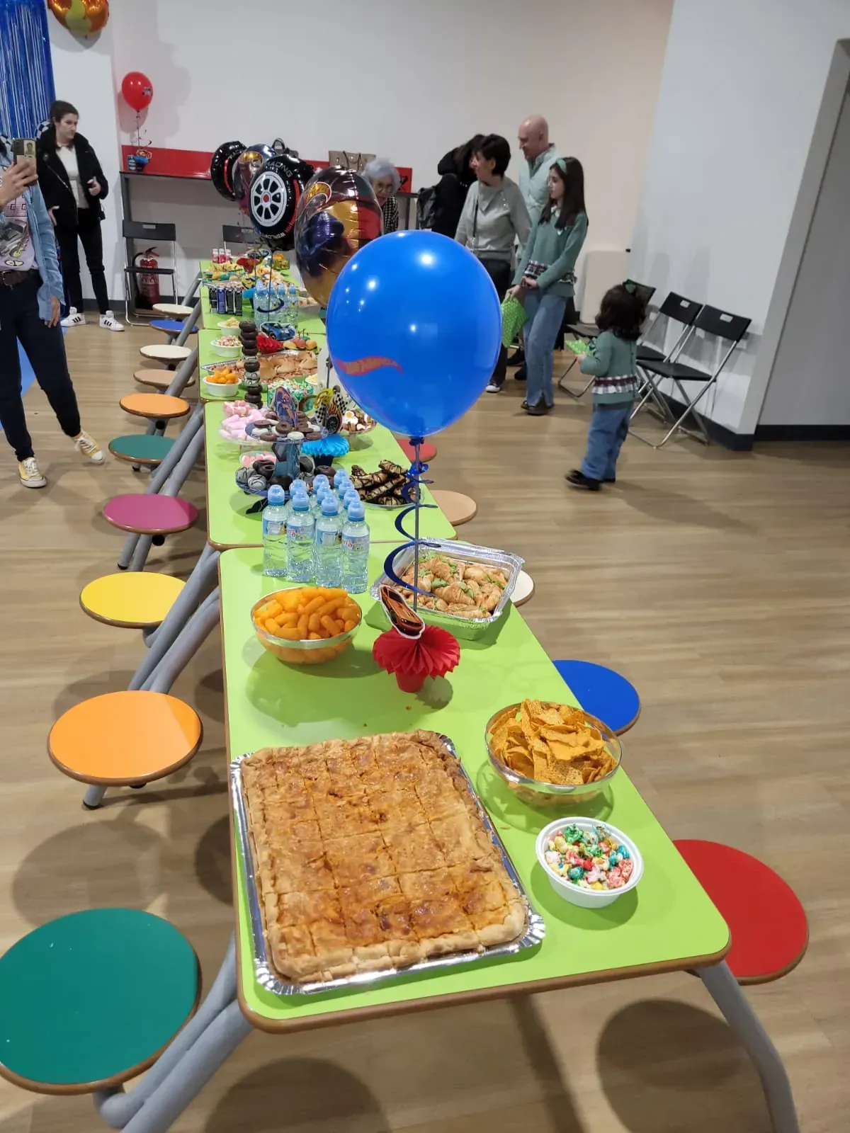 Mesa de fiesta con bocadillos, globo azul y gente en una habitación.