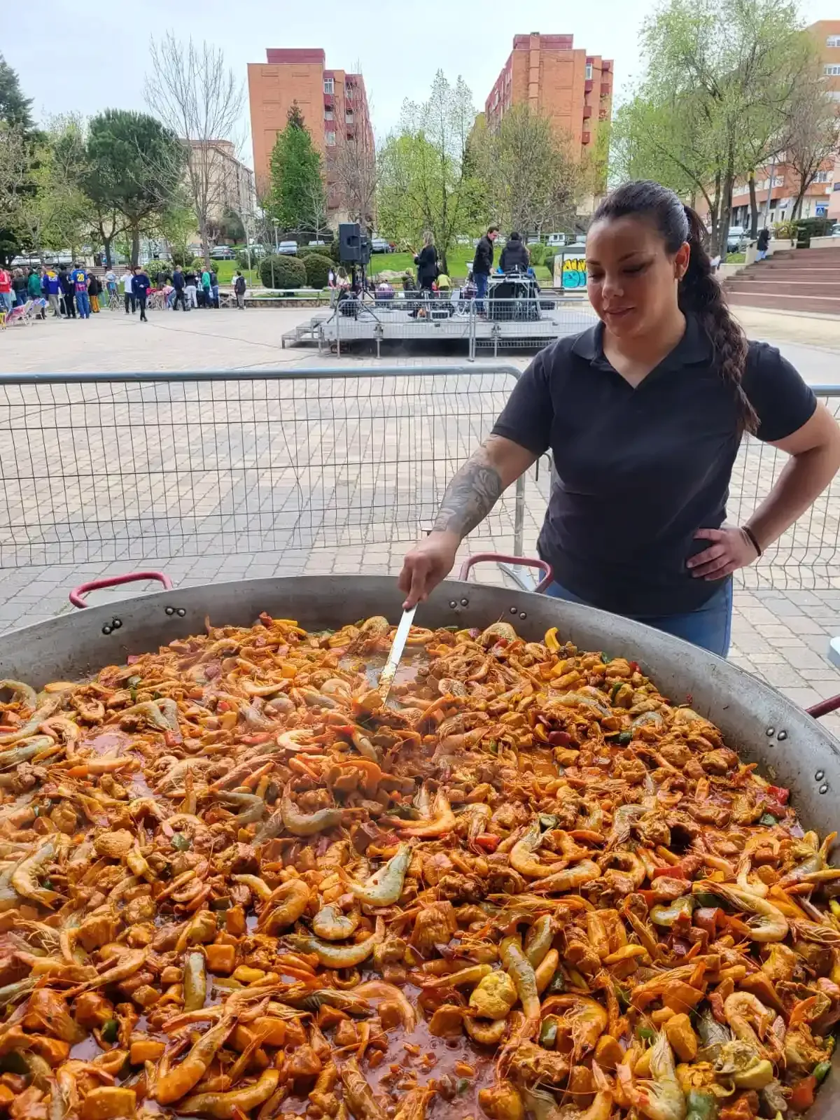 Mujer revolviendo paella en una sartén grande al aire libre. Gente al fondo, cerca del escenario.