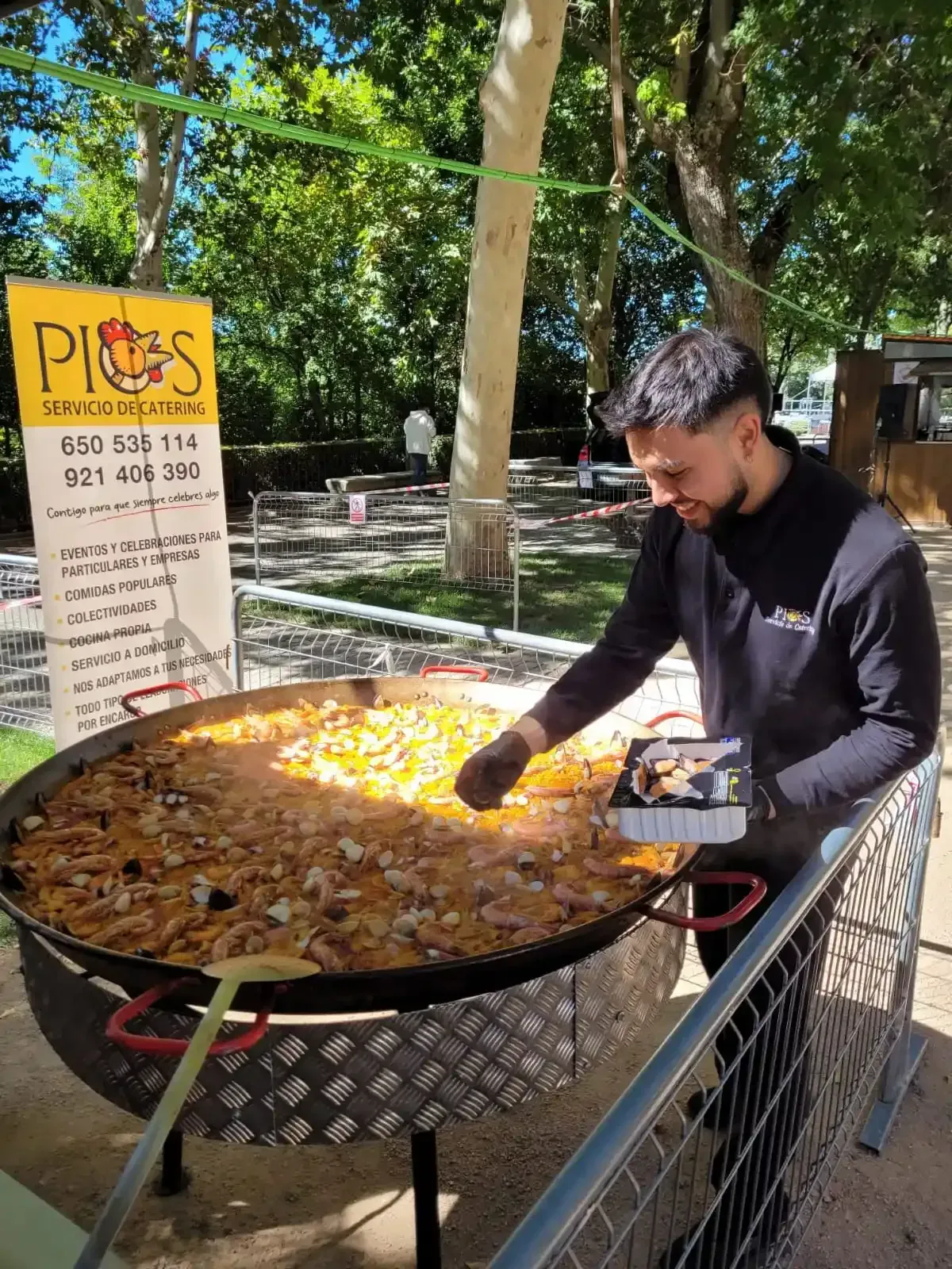 Hombre cocinando paella en una sartén grande al aire libre. Cartel visible con información de contacto.