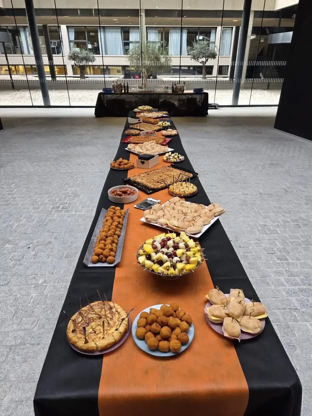 Una mesa larga repleta de comida variada, incluidos pasteles, albóndigas y sándwiches, en un edificio moderno.