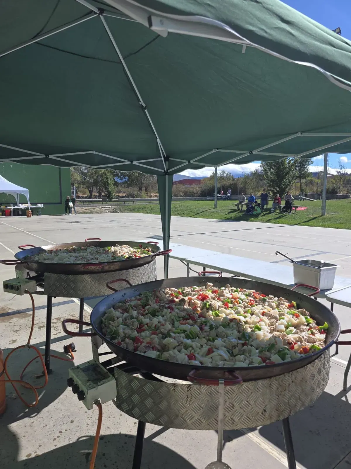 Dos grandes paelleras cocinándose bajo una carpa verde en un evento al aire libre.