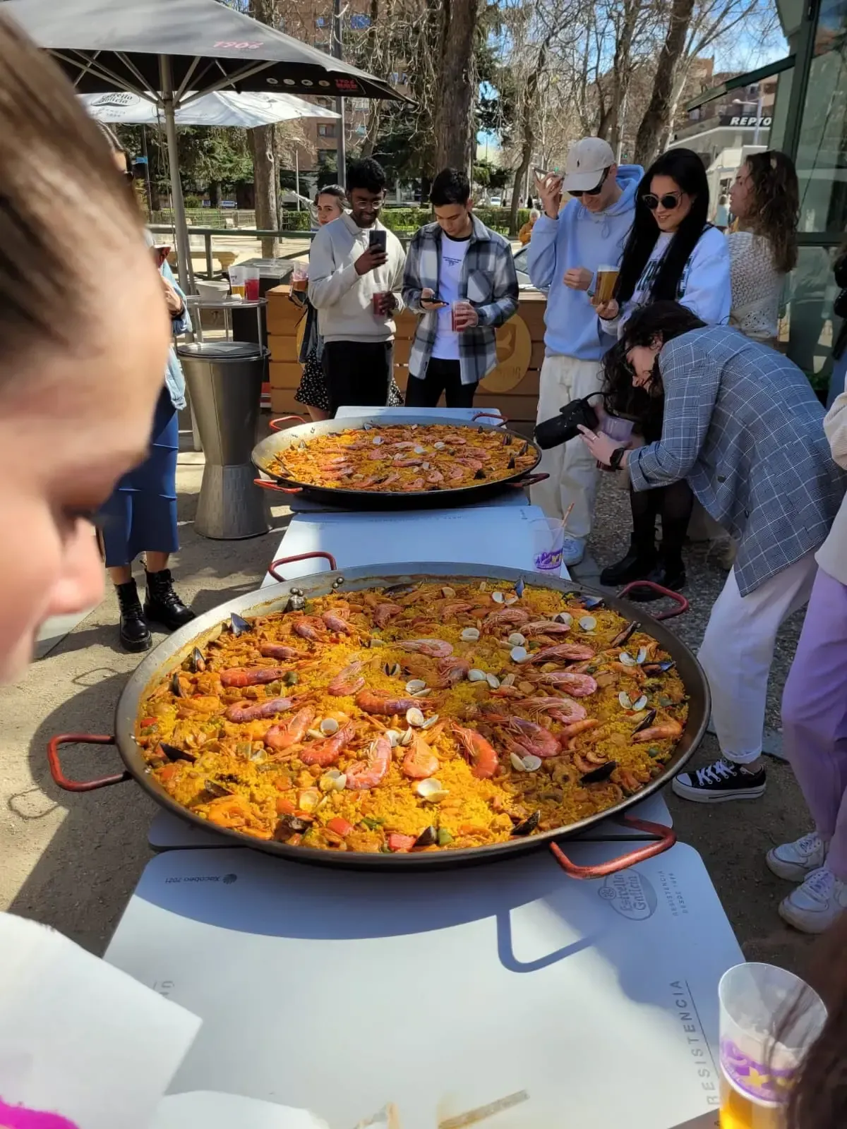 Dos grandes paelleras sobre una mesa, rodeadas de gente al aire libre en un día soleado.
