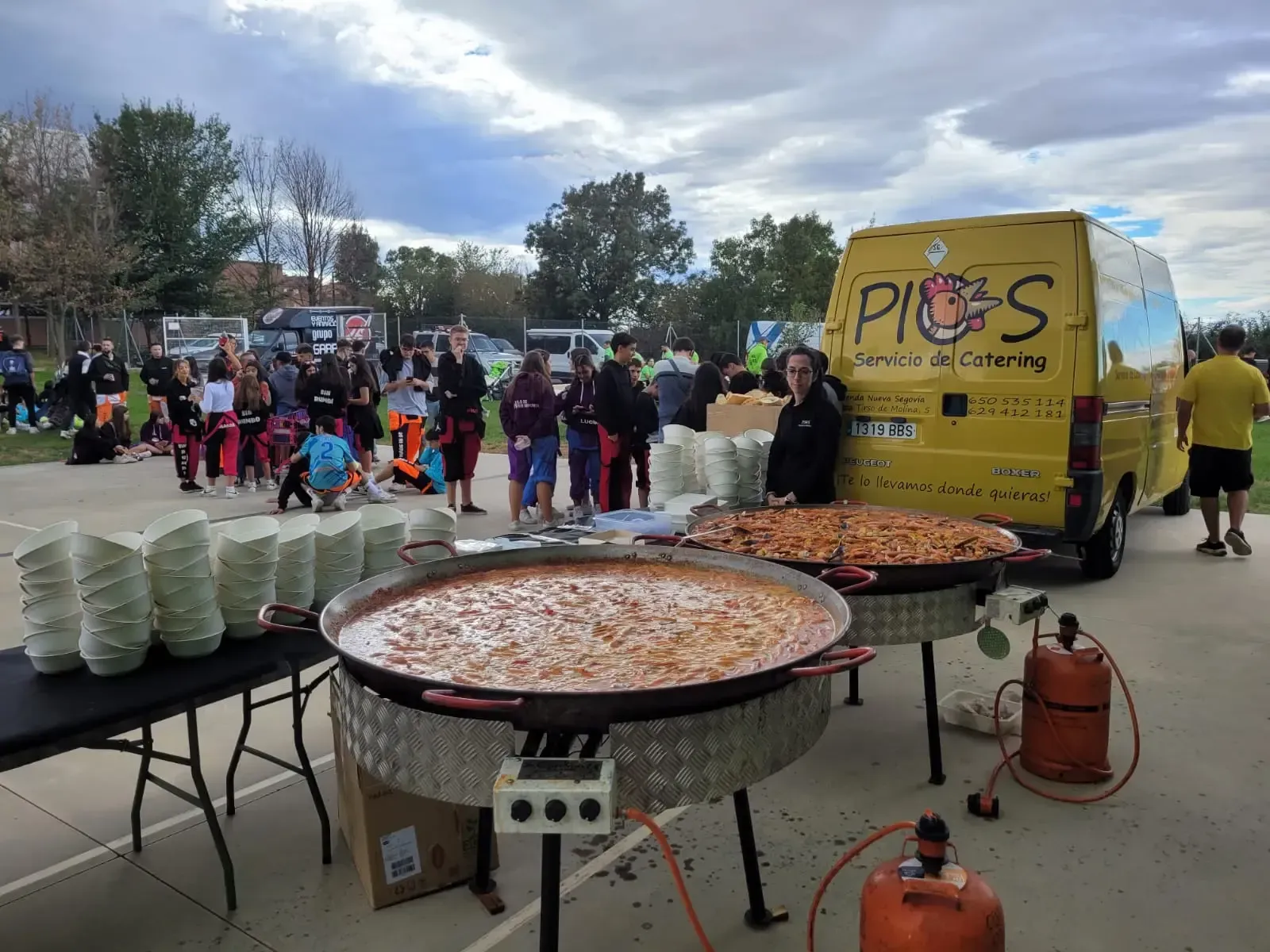 Grandes paelleras con servicio de comida desde una furgoneta amarilla; gente esperando.