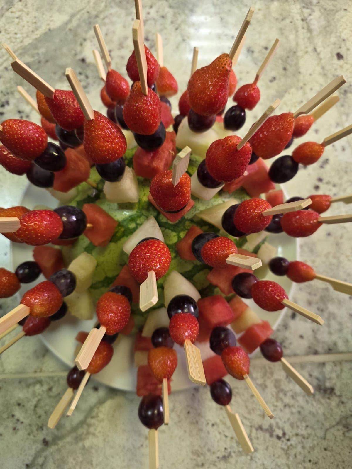 Brochetas de frutas dispuestas como centro de mesa colorido: fresas, uvas, melón en palitos de madera, plato blanco.