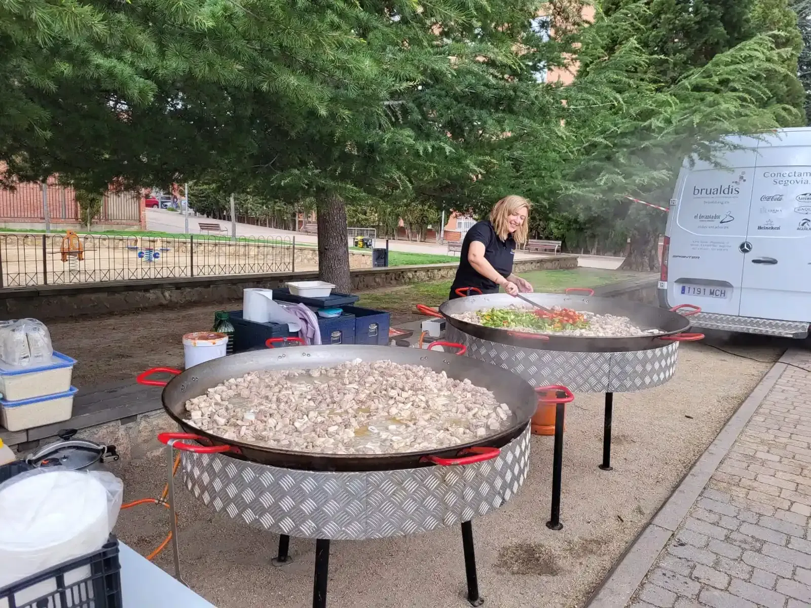 Mujer cocinando paella en grandes sartenes al aire libre, cerca de una camioneta blanca y árboles.