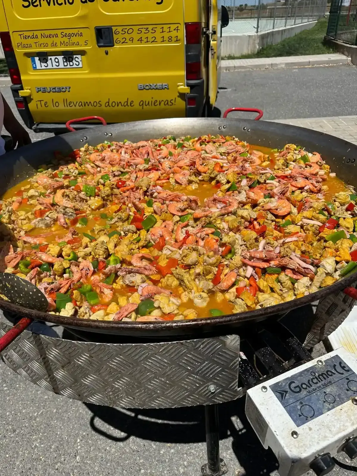 Gran paella con camarones en una sartén gigante sobre un quemador al aire libre, cerca de una furgoneta amarilla.