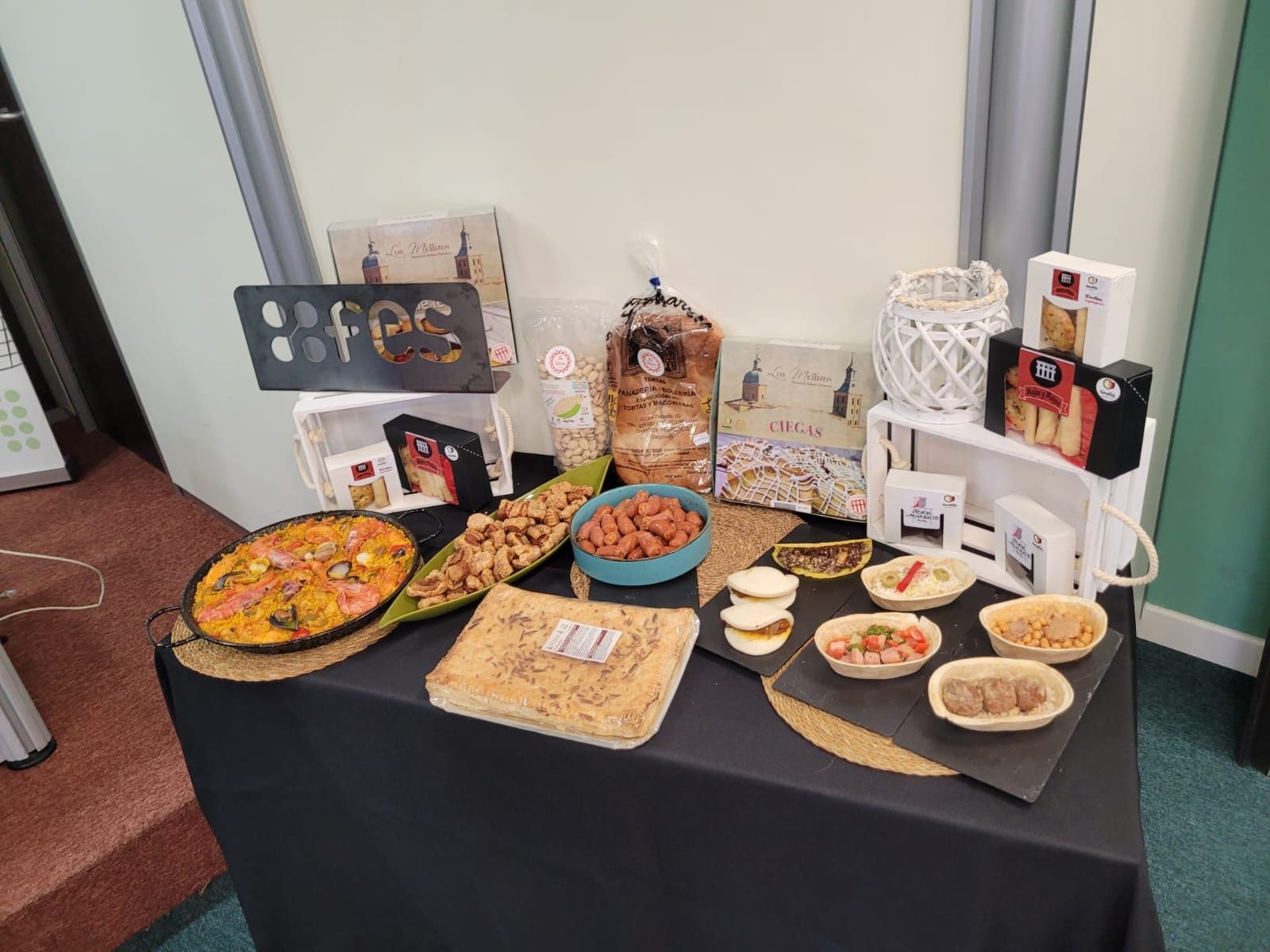 Mesa de tapas españolas. Diversos platos, galletas y cajas dispuestas sobre un mantel negro en una feria comercial.