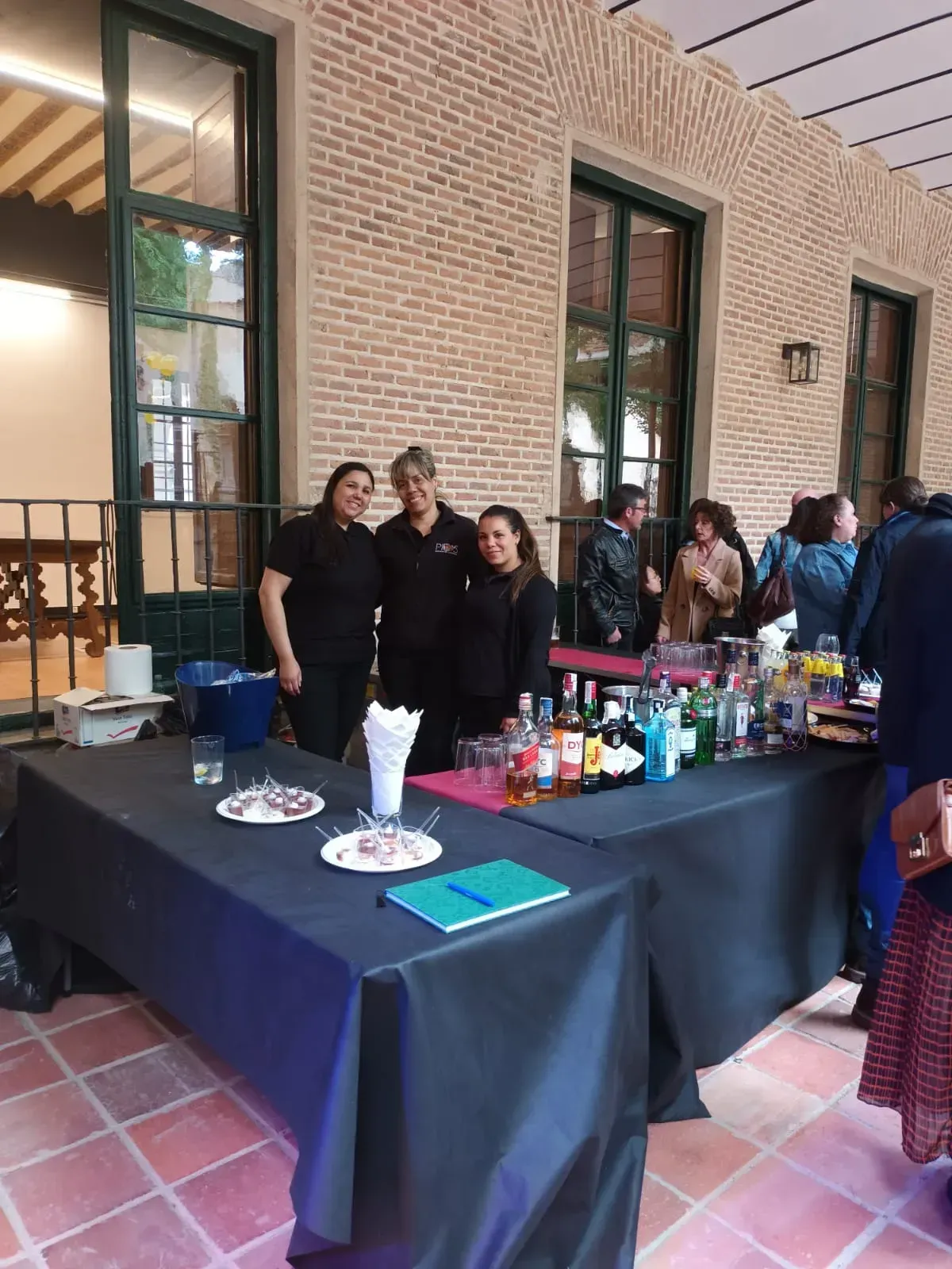 Tres personas detrás de una barra sirviendo bebidas en un evento, frente a una pared de ladrillos.