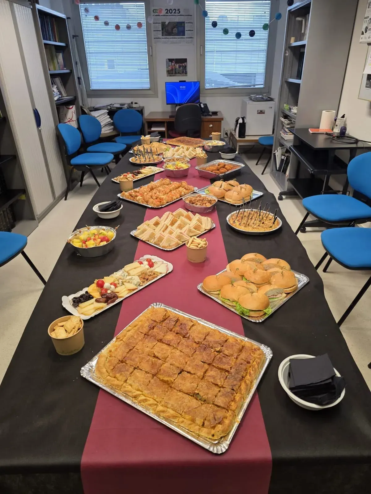 Una mesa larga repleta de diversos platos preparados. Sillas azules rodean la mesa en un ambiente de biblioteca.