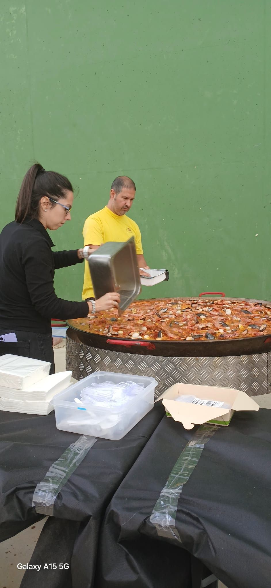 Dos personas sirviendo comida de una paellera grande. Fondo de pared verde.