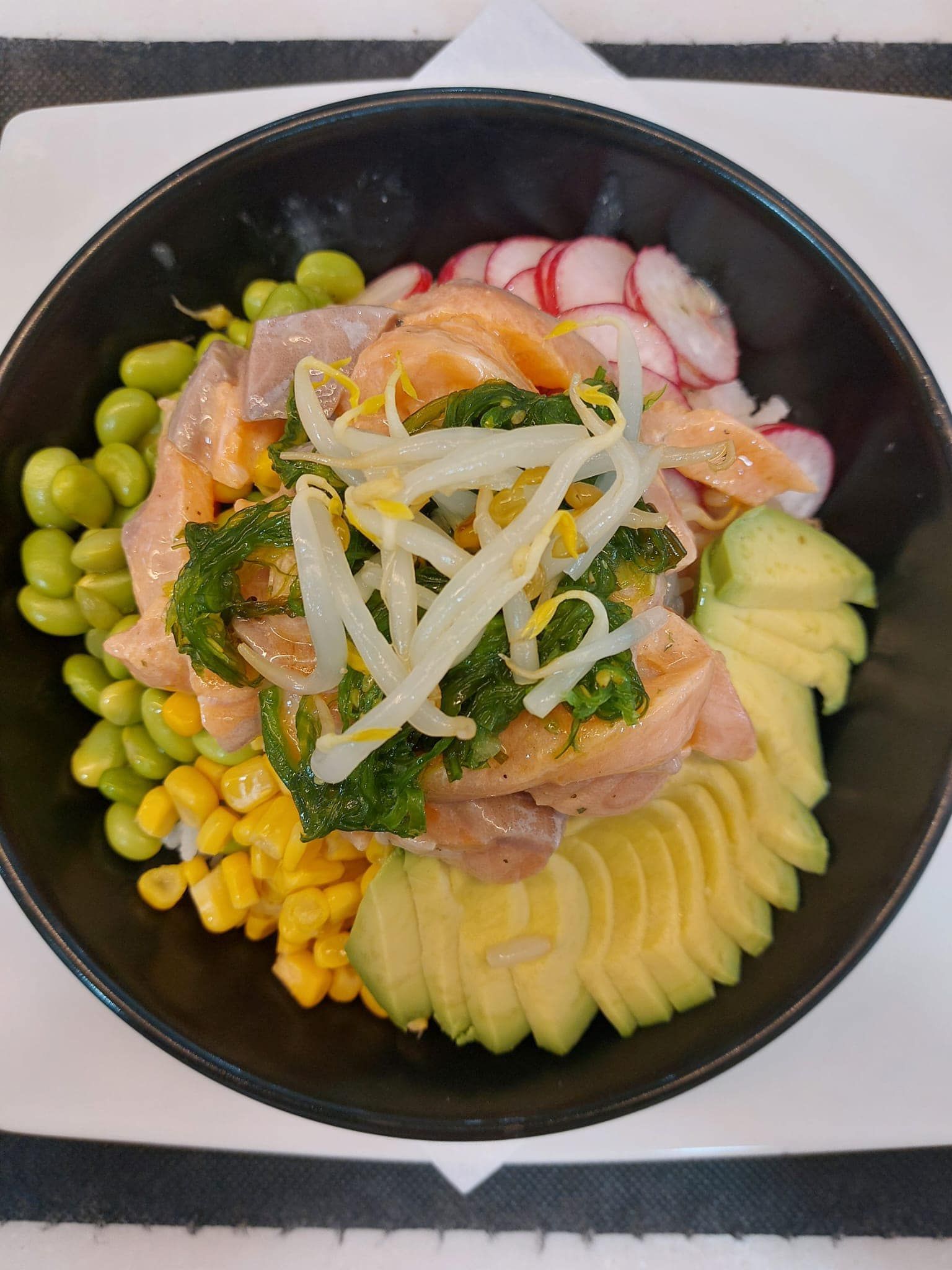 Un tazón de comida con aguacate, maíz y rábanos en una mesa.