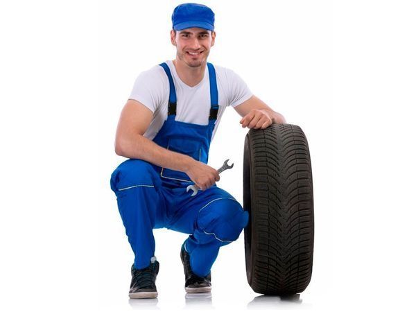 Un hombre con un mono azul está arrodillado junto a un neumático y sostiene una llave inglesa.