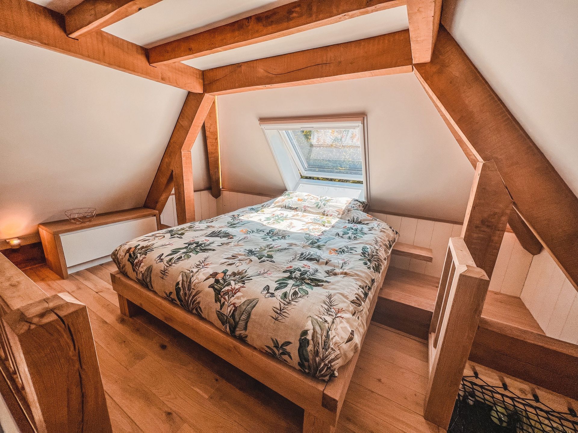 Slaapkamer met houten balken, dakraam boven het bed, beddengoed met bloemenprint en houten vloer.
