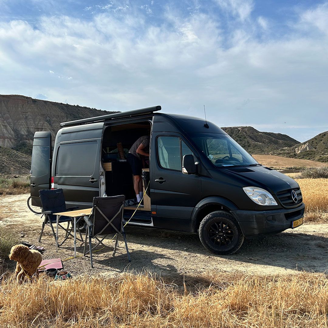 Zwarte bestelwagen met open deur, campingstoelen en een hond in een droge buitenomgeving.