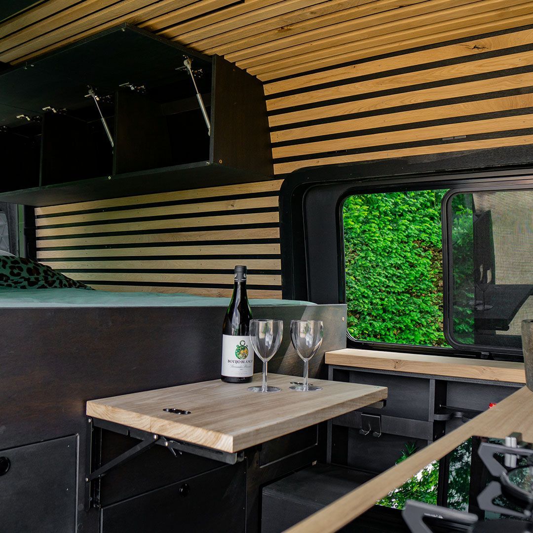 Gezellig interieur van de camper: houten lambrisering, een klein tafeltje met wijn, groen zichtbaar door een raam.
