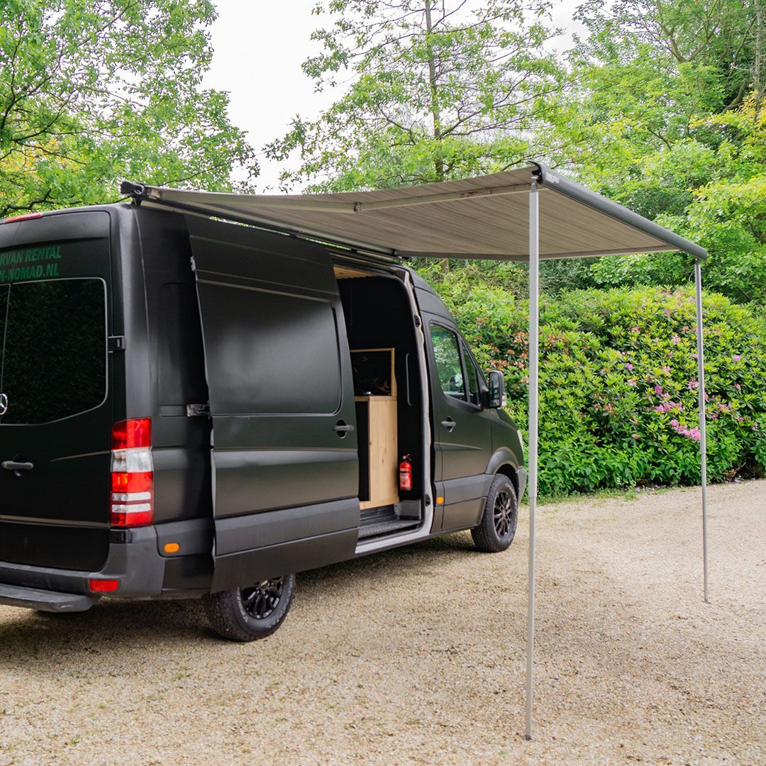 Zwarte bestelwagen met uitgeschoven luifel, geparkeerd op grind. Deur open, bomen op de achtergrond.