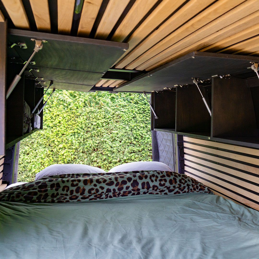 Interieur van een camperbusje met een bed, open opbergruimte bovenin en uitzicht op groen gebladerte.