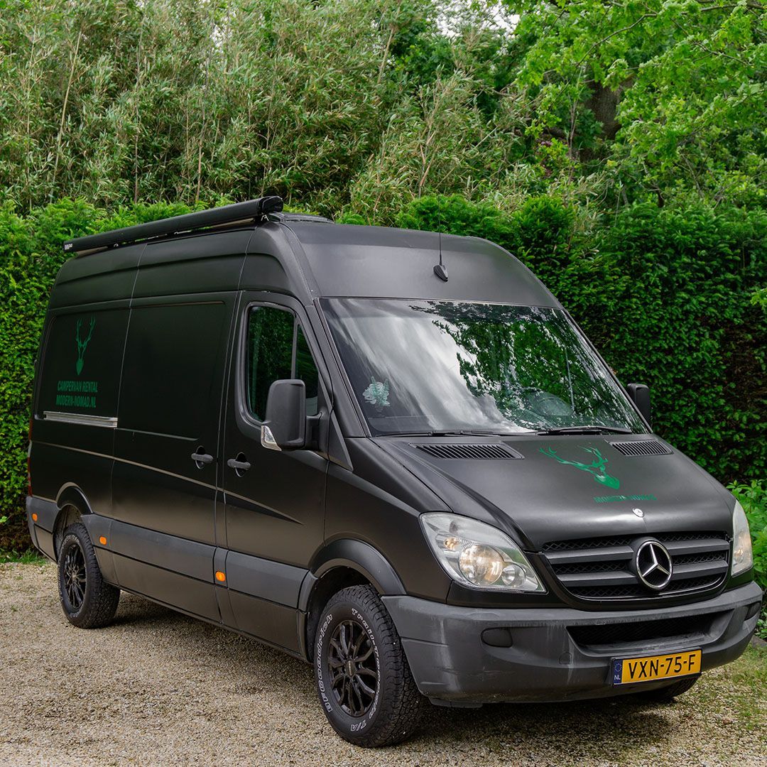 Zwarte Mercedes-bus met imperiaal geparkeerd voor een groenstrook; groen logo op de zijkant en motorkap; Nederlands kenteken.