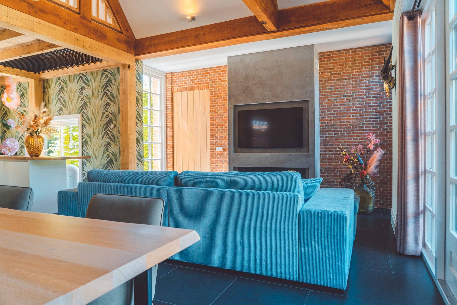 Woonkamer met een turquoise corduroy bank, bakstenen muur, open haard, houten balken en grote ramen.