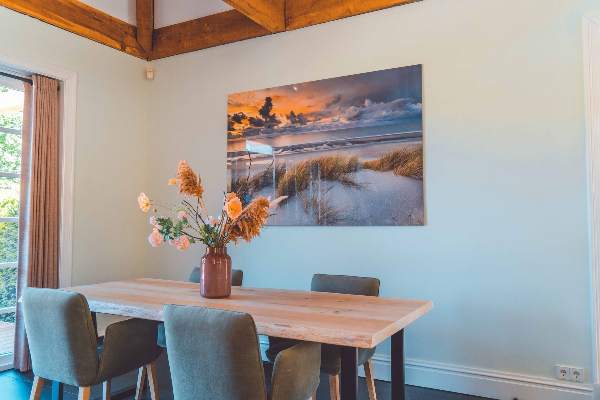 Eetkamer met houten tafel, grijze stoelen, bloemstuk en een kunstwerk met een strandtafereel aan de muur.