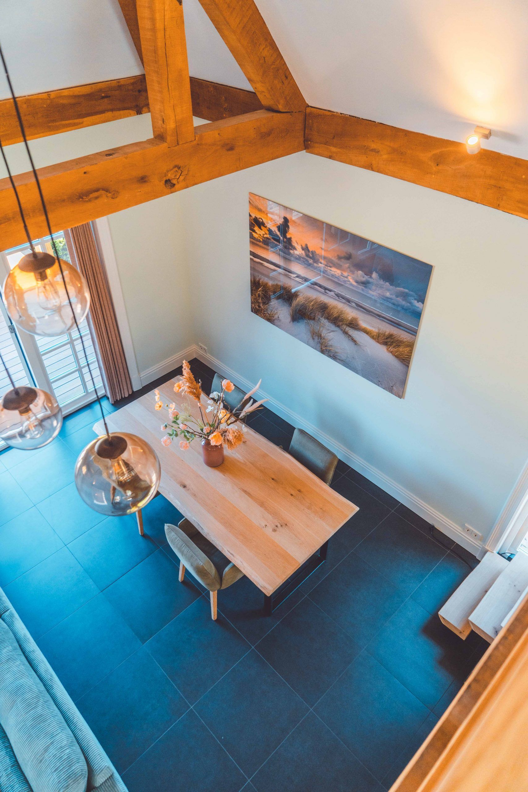 Een opname vanuit een hoog perspectief van een eetkamer met houten balken, een grote tafel en bolvormige hanglampen. Blauwe vloer.