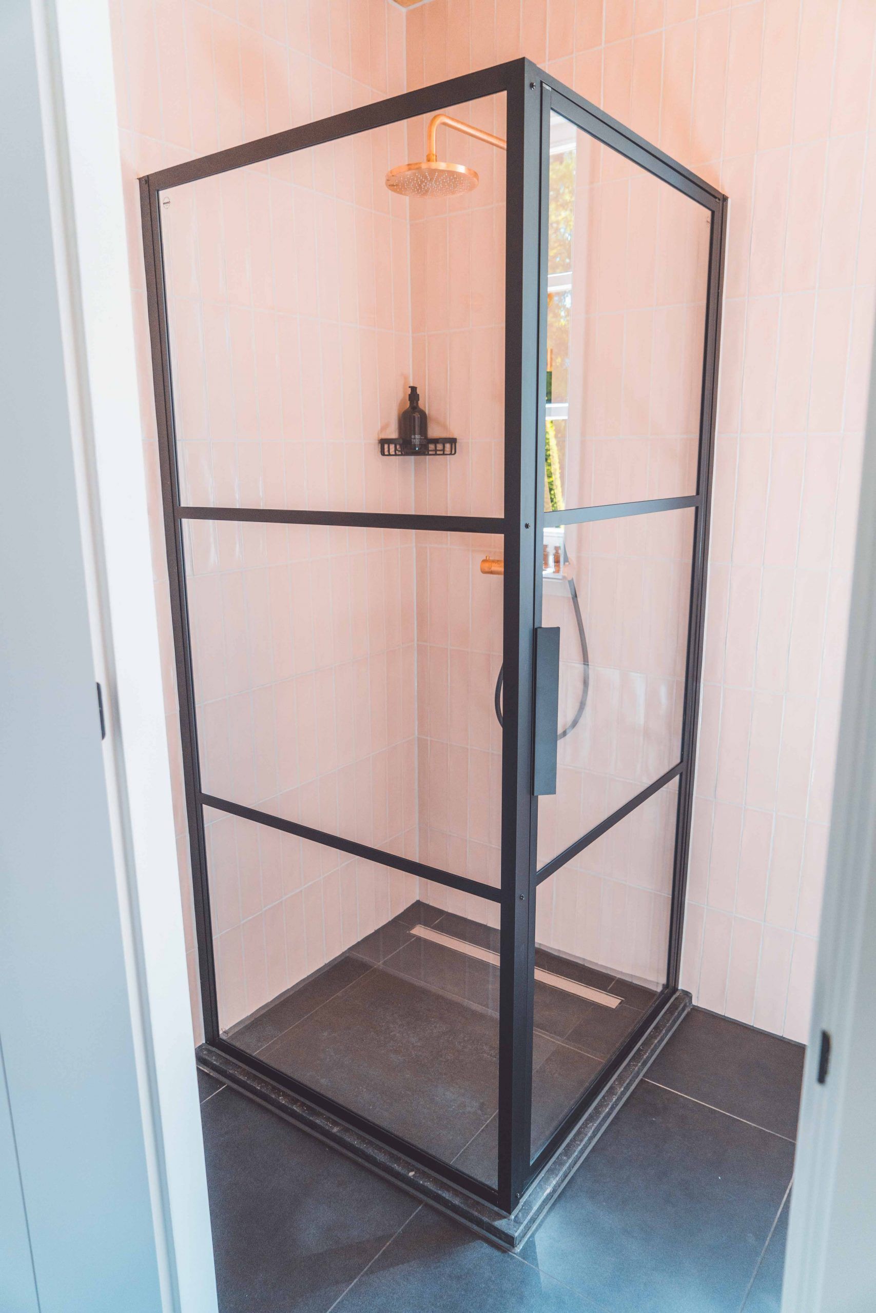 Douchecabine met zwart frame en glazen panelen in de hoek van een badkamer met roze tegels. Douchekop en plankje met producten zichtbaar.