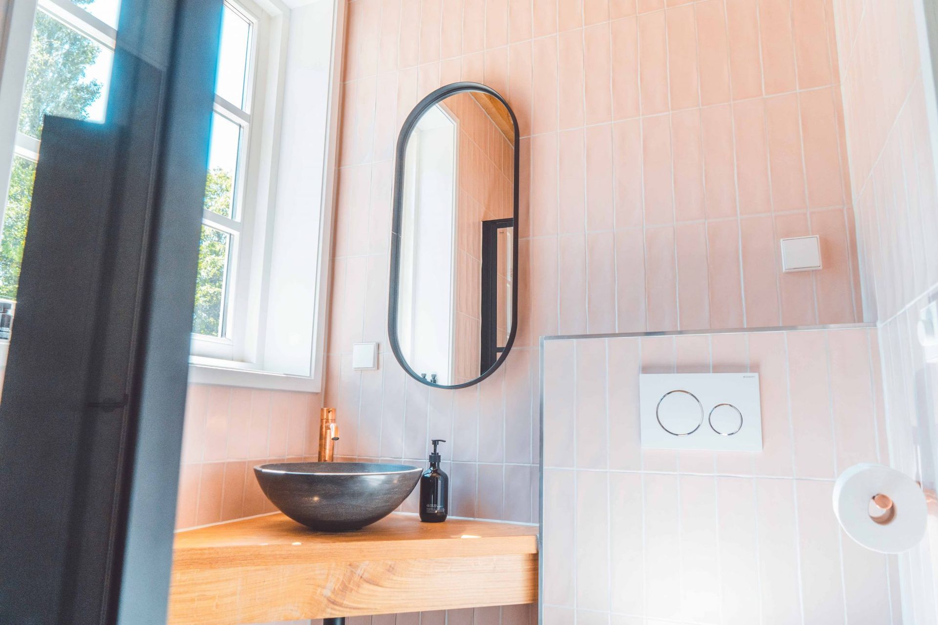 Badkamer met roze betegelde muren, ovale spiegel, donkere wastafel, houten aanrechtblad en raam.