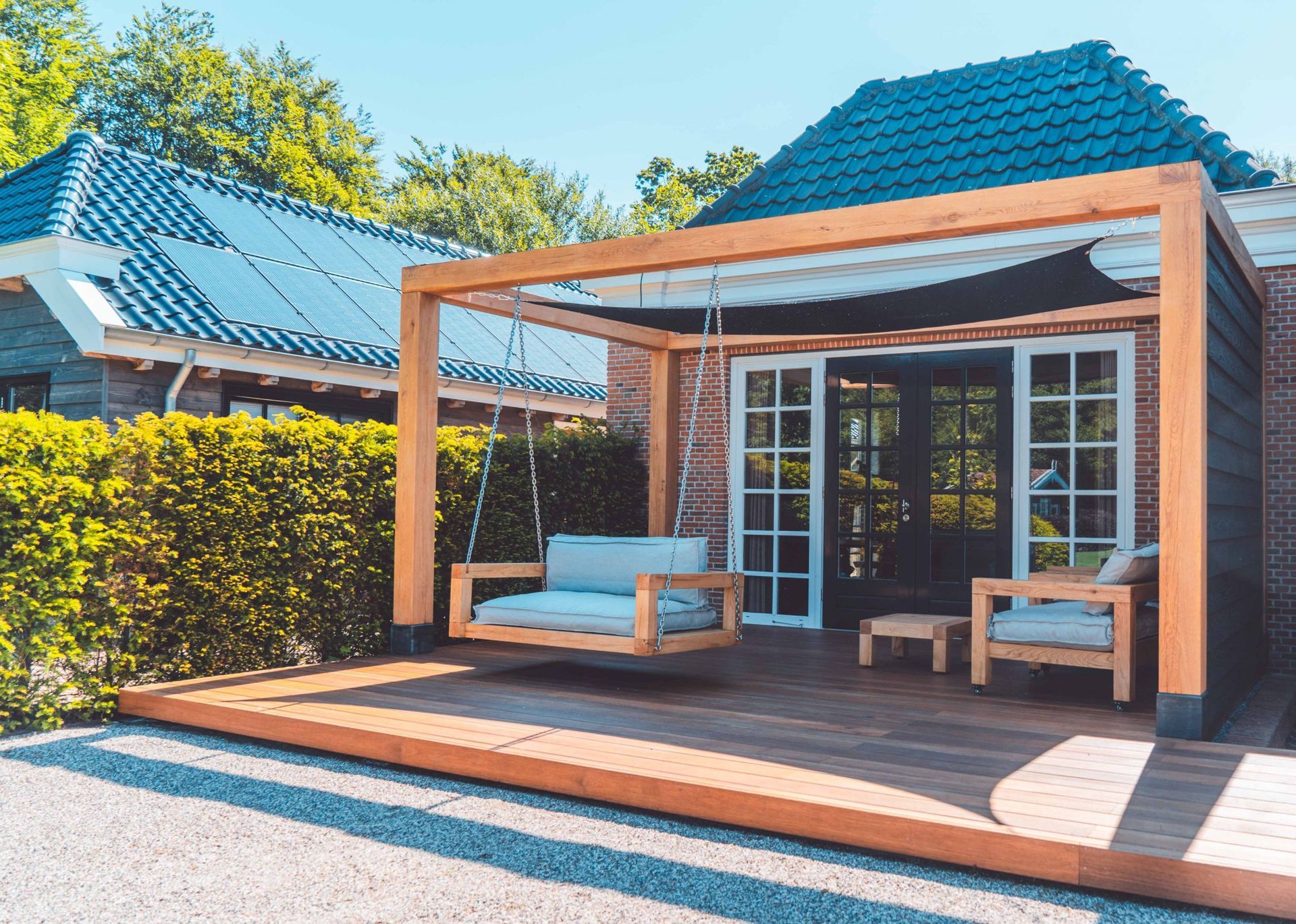 Houten terras met een schommel, zitgedeelte en een bijgebouw met openslaande deuren. Zonnige dag.