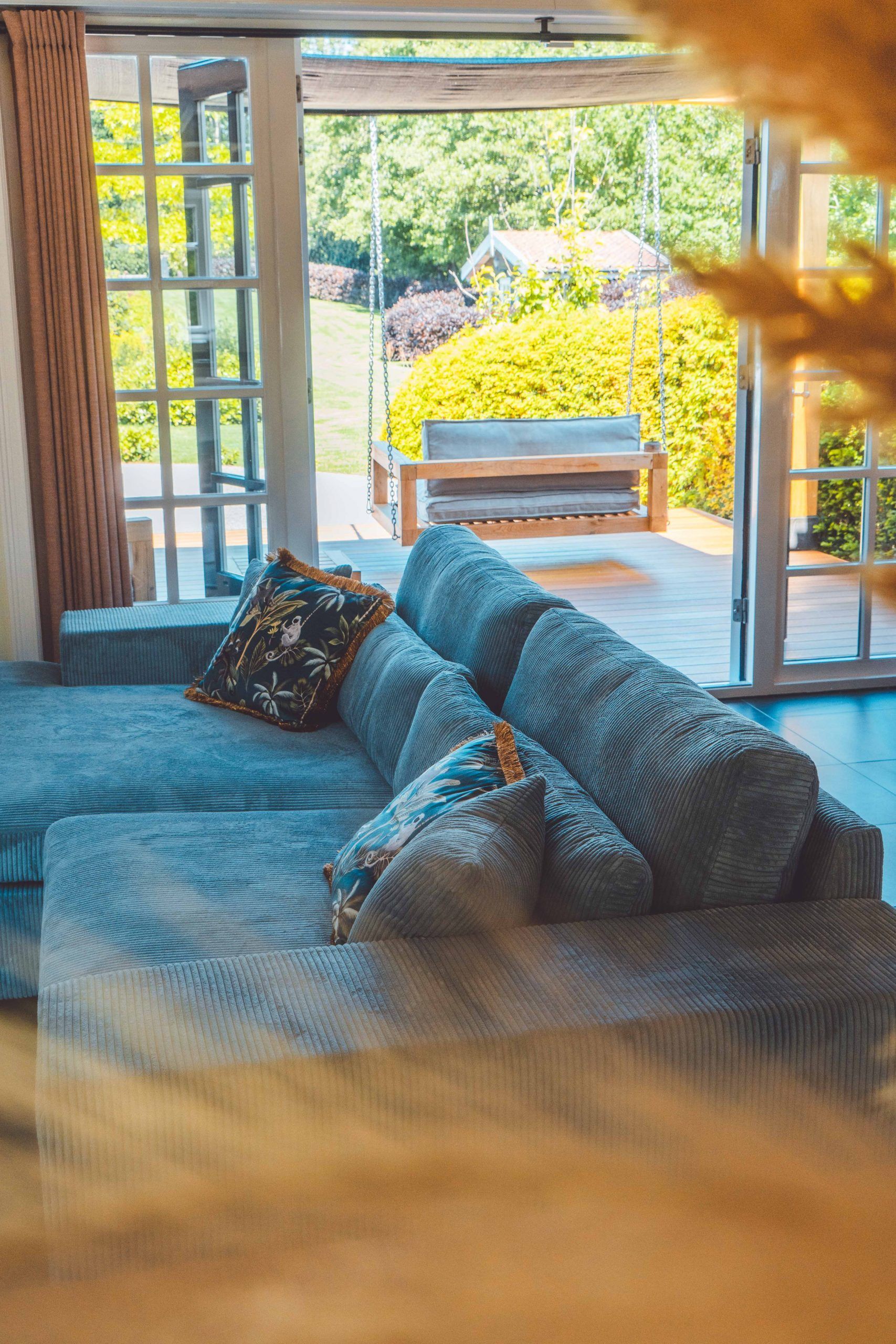 Een blauwe hoekbank staat tegenover een open deur naar een terras met een houten schommel en planten.
