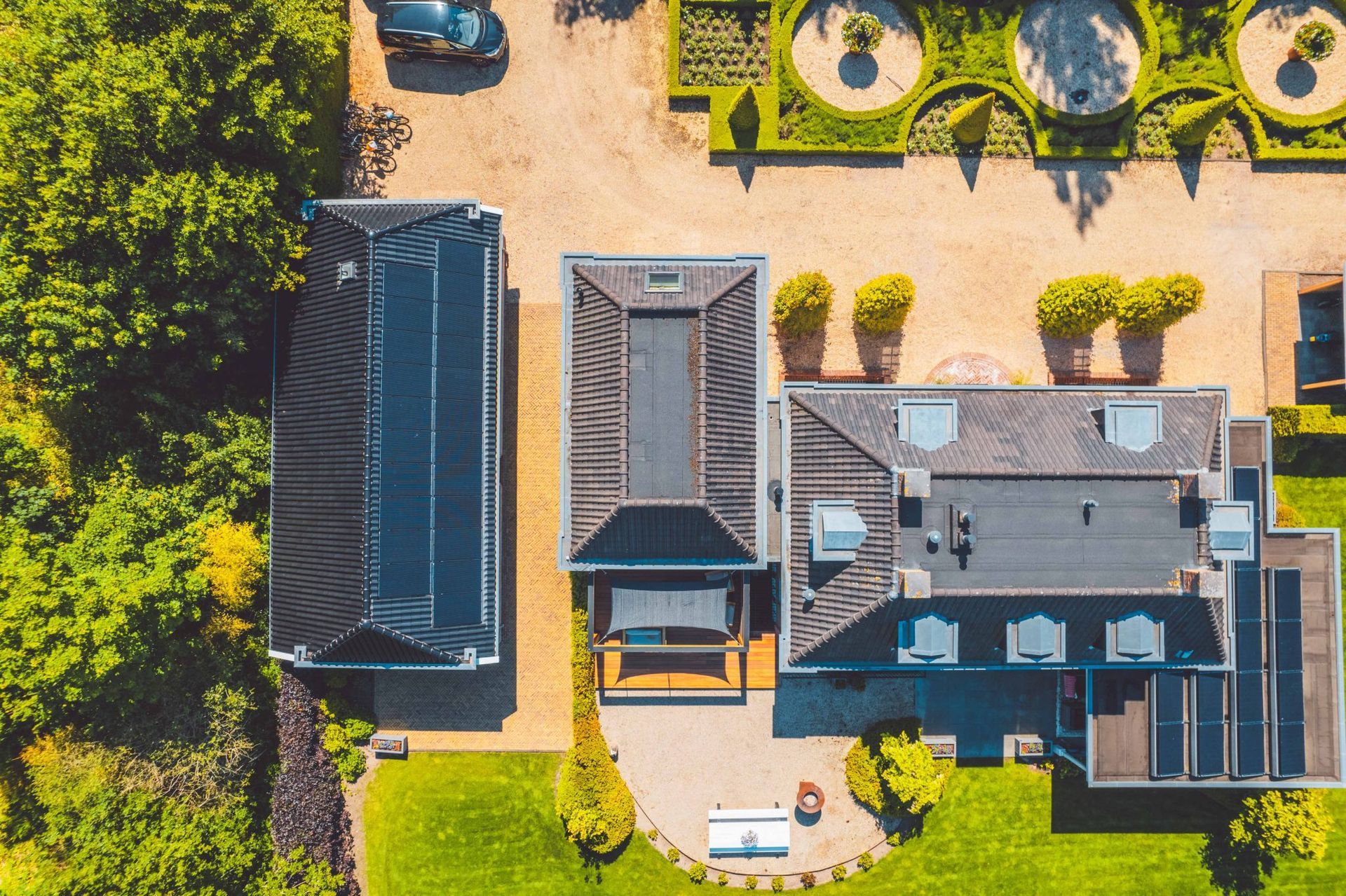 Luchtfoto van een complex met meerdere gebouwen, weelderige tuinen en een grindpad; er staat een auto geparkeerd op het pad.