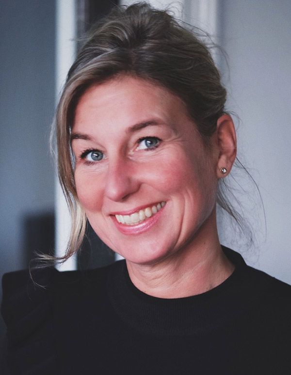 Vrouw met blond haar, lachend, draagt een zwarte top. Binnen, neutrale achtergrond.