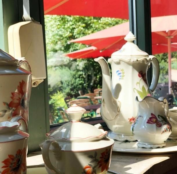 Ein Porzellan-Teeservice steht auf einem Tisch an einem sonnigen Caféfenster, draußen stehen rote Sonnenschirme.
