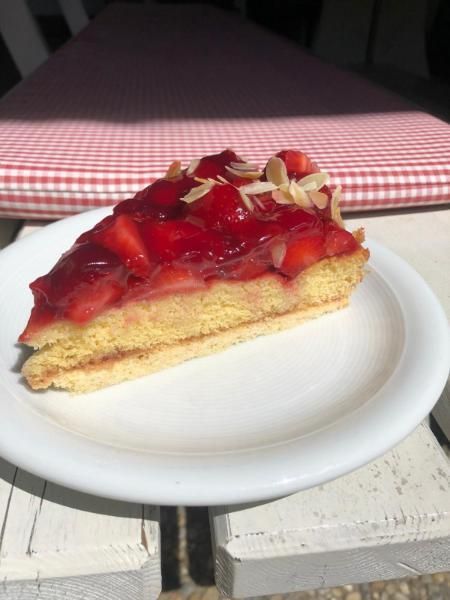 Ein Stück Erdbeertorte auf einem weißen Teller auf einem Picknicktisch vor einem rot-weiß karierten Tischtuch.