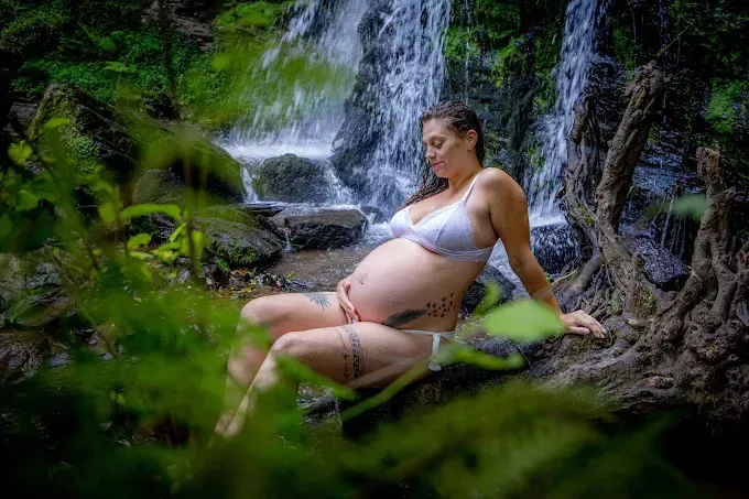 Una mujer embarazada sentada en un tronco frente a una cascada, rodeada de exuberante vegetación.