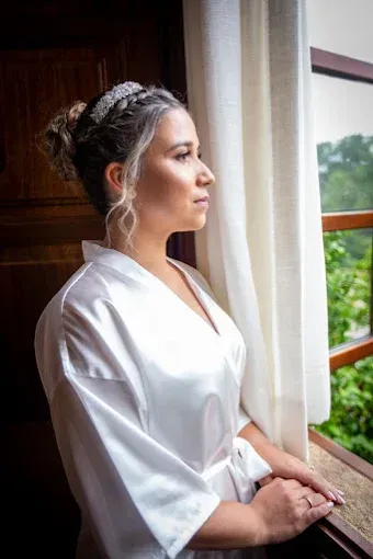 Una persona vestida con una bata de satén blanco y un peinado trenzado mira por la ventana.