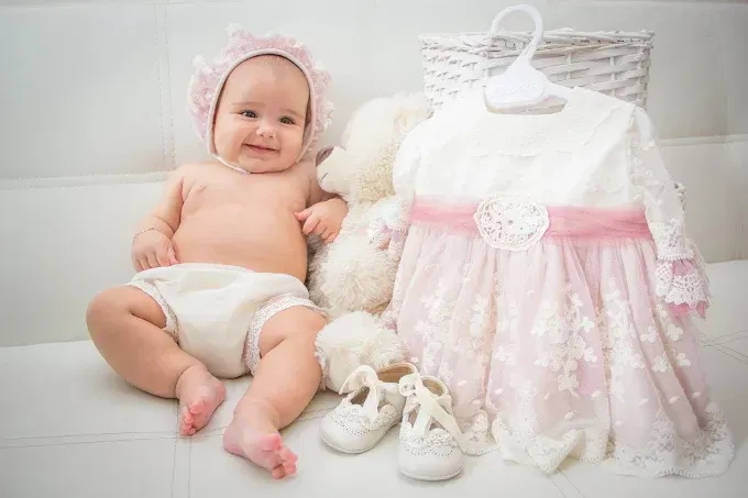 Bebé sonriendo junto a un vestido blanco y rosa, un osito de peluche y zapatos sobre una suave manta blanca.