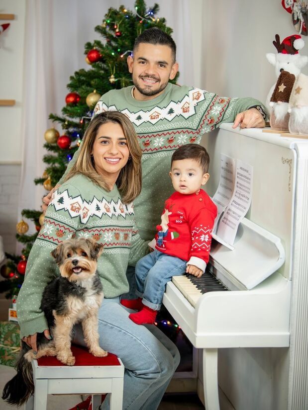 Familia con suéteres navideños a juego posando con un niño pequeño, un perro y un piano blanco junto a un árbol decorado.