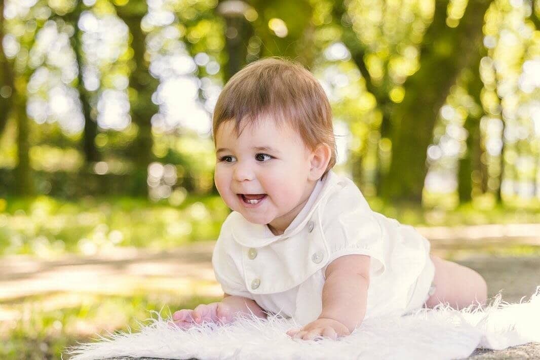 Un bebé sonriente, vestido de blanco, tumbado sobre una manta blanca texturizada en un parque soleado.
