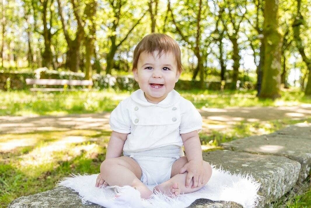 Un bebé sonriente está sentado sobre una manta blanca en un muro de piedra en un parque soleado y lleno de árboles.