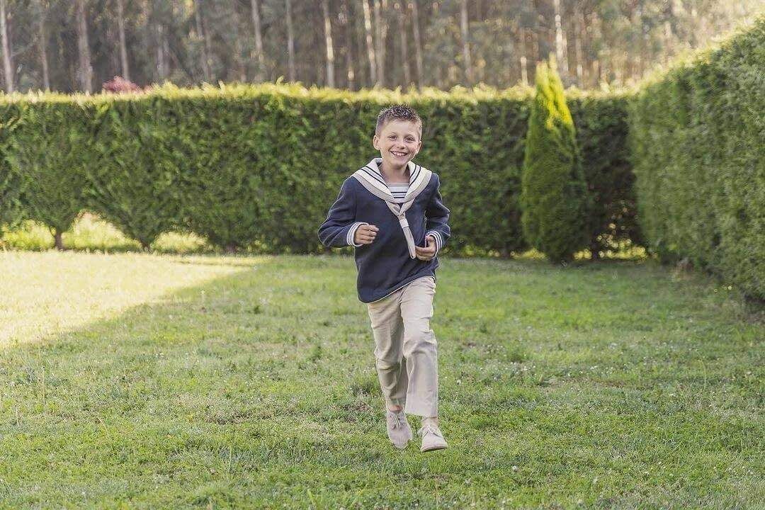 Un niño sonriente corre por un césped frente a un seto alto.