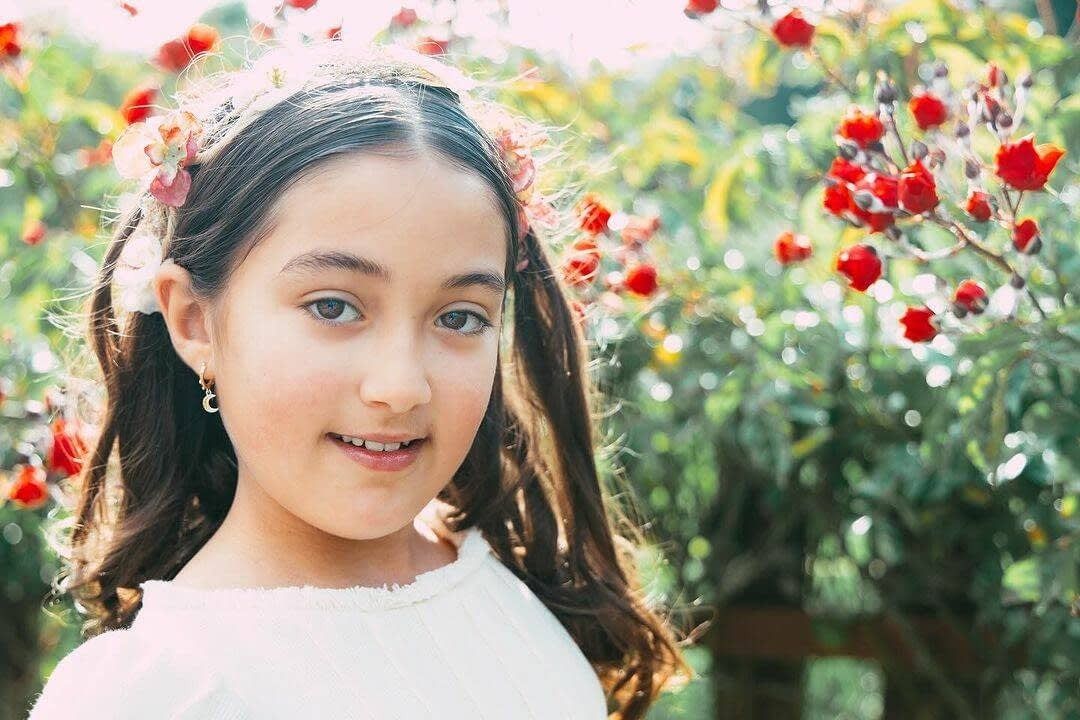 Una persona con una diadema floral sonríe dulcemente frente a un rosal en flor.
