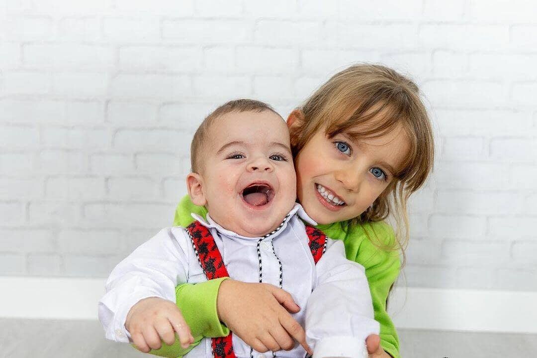 Un niño sonriente sostiene a un bebé feliz frente a un fondo de ladrillos blancos.