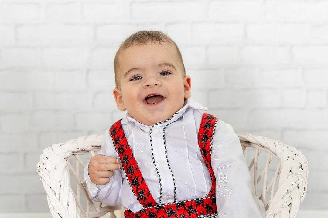 Un niño sonriente, con una camisa blanca y tirantes estampados en rojo y negro, sentado en una silla de mimbre blanca.