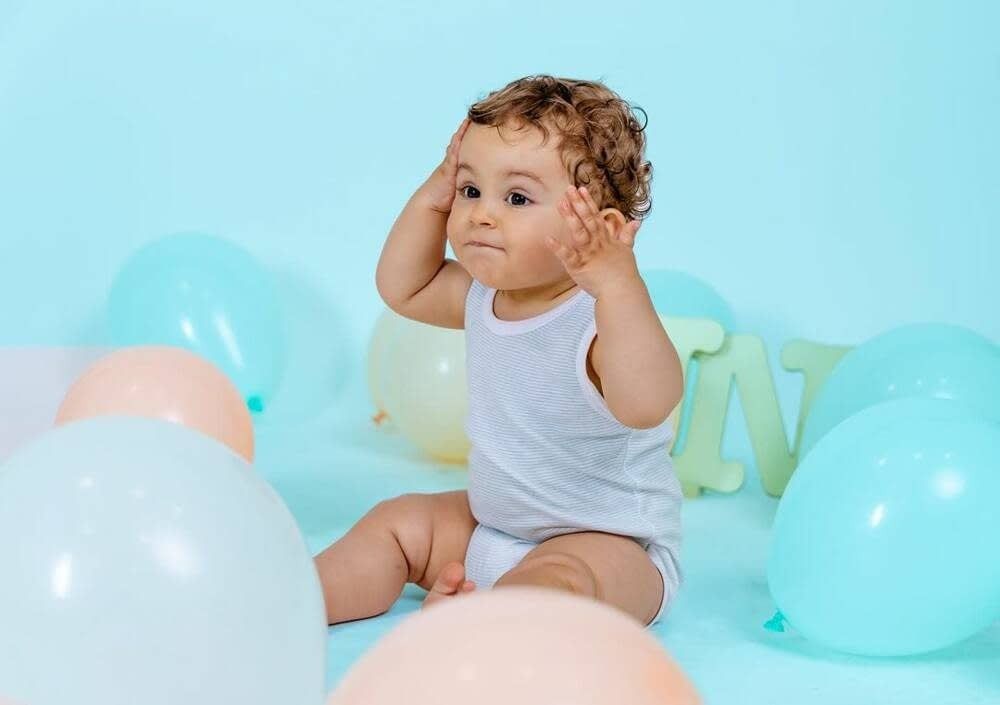 Un bebé con cabello rizado está sentado en un suelo azul rodeado de globos azul 