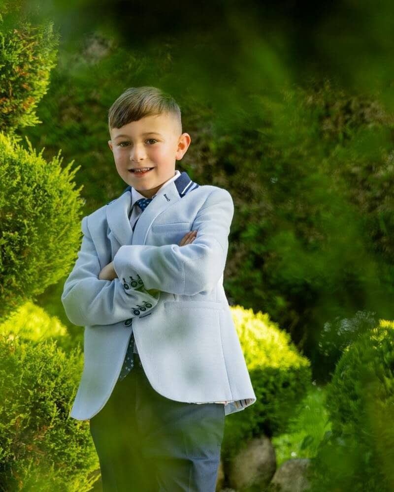 Un niño sonriente con los brazos cruzados, vestido con una chaqueta azul claro, de pie al aire libre entre arbustos verdes.