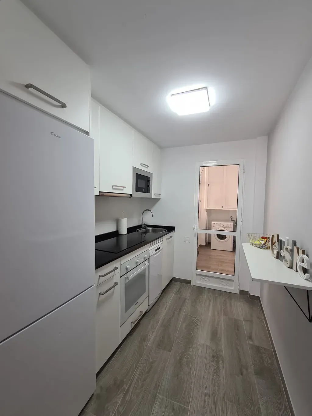 Pequeña cocina blanca con nevera, armarios, electrodomésticos y suelo gris. Una puerta da acceso a la zona de lavandería.