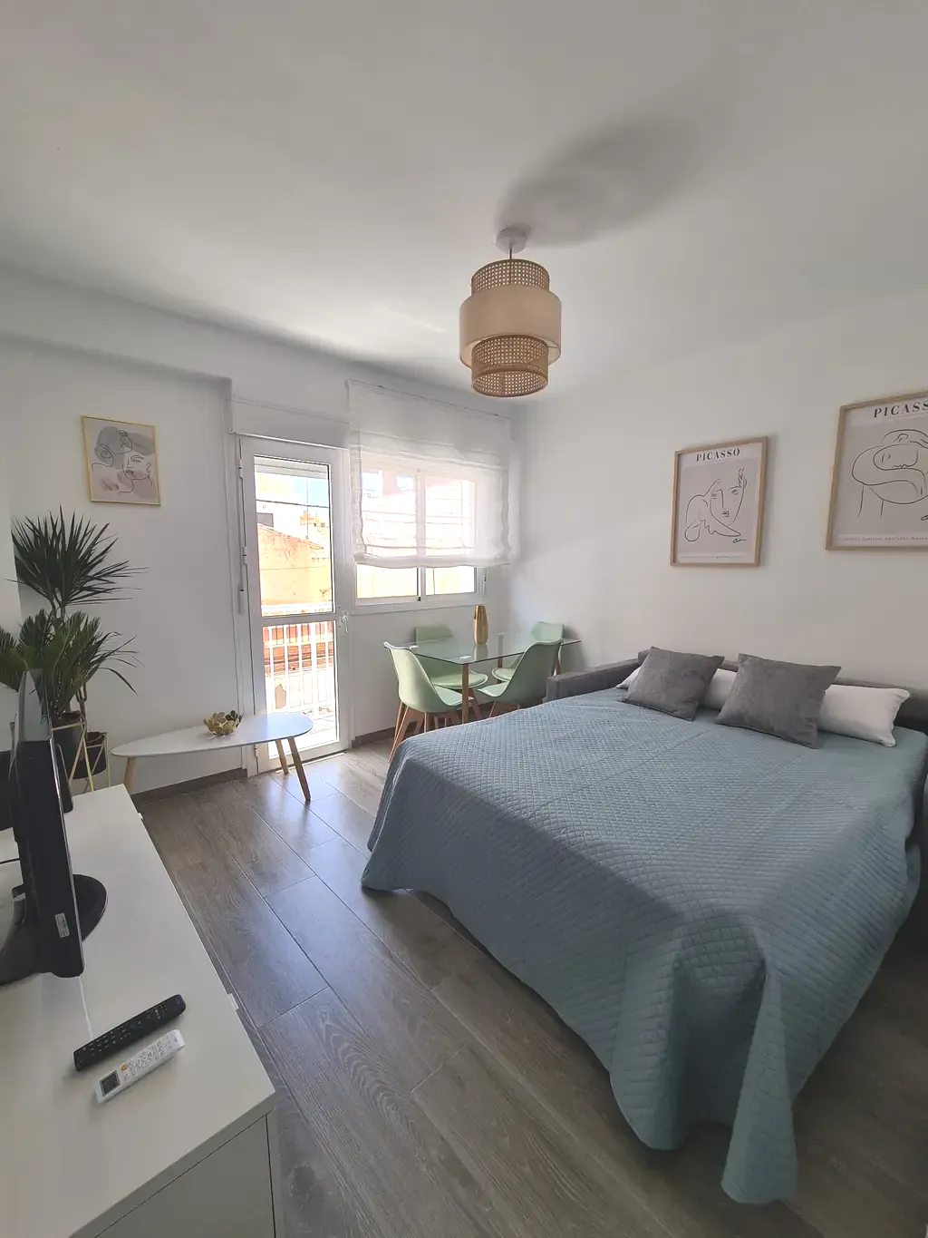 Dormitorio con colcha azul, mesita, sillas verdes y plantas; luminoso con paredes blancas.