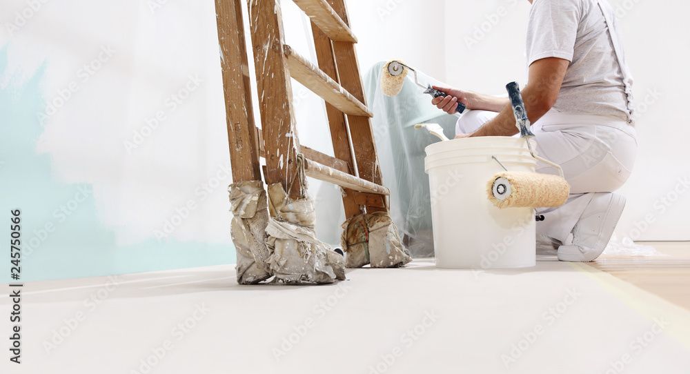 Maler auf dem Boden neben Leiter und Farbeimer, streicht Wand blau, trägt Weiß.